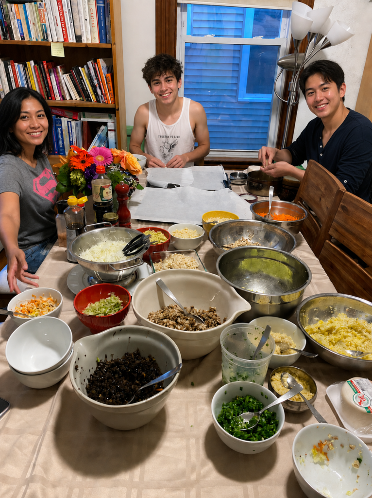 Three smiling people sit around a dining table covered with bowls of chopped vegetables and other ingredients, appearing to prepare a meal together. A vase of colorful flowers sits near the center of the table, with bookshelves and a window in the background.
